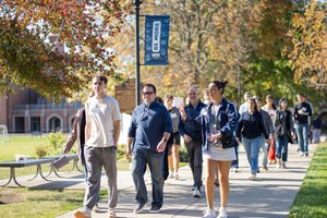 A diverse group of students and adults walks a sunny university path in autumn, with a student guide gesturing and speaking.