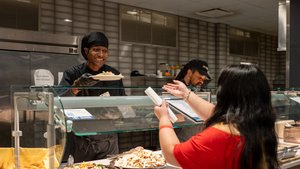 Two smiling workers serve students a variety of food options at a buffet line.