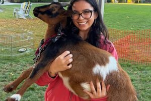 A smiling woman wearing glasses and a red jacket holds a brown and white goat in a grassy outdoor area.