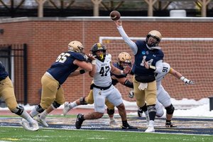 A football player in a blue jersey throws a pass while being pursued by opponents in white jerseys on a field.