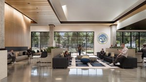 People sit and chat in a modern lounge area with large windows showing trees and a university seal on the wall.