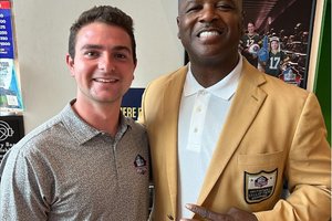 Two men smile at the camera, one wearing a gray polo and the other a gold jacket with a Hall of Fame patch.