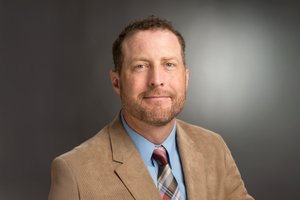 A man with short curly hair and a beard wears a tan blazer, blue shirt, and striped tie against a gray background.