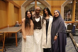 Four women stand closely together inside a room with wooden walls and large windows, smiling at the camera.