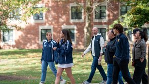 A group of seven people walk together on a sidewalk beside a grassy area with a brick building in the background.