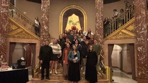 A group of people pose on a grand staircase with ornate marble columns and a large plant in a red vase behind them.