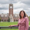 A woman in a pink blazer smiles while leaning on a railing with a large brick building and green lawn behind her.
