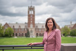 A woman in a pink blazer smiles while leaning on a railing with a large brick building and green lawn behind her.