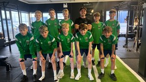 A group of ten boys in green sportswear and one man in a black shirt pose together inside a gym with exercise equipment.