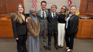 Six young adults dressed in business attire stand arm in arm smiling in a courtroom with flags behind them.