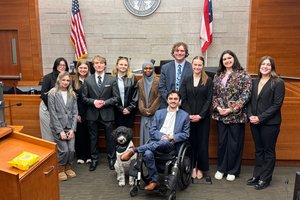 A group of ten people and a black-and-white dog pose together in a courtroom with flags and a seal on the wall behind them.