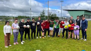A group of people stand closely together on a grassy sports field holding Gaelic footballs under a partly cloudy sky.