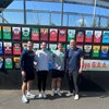 Four people stand side by side smiling in front of a wall displaying colorful GAA club crests under a clear blue sky.
