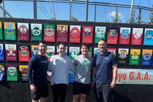 Four people stand side by side smiling in front of a wall displaying colorful GAA club crests under a clear blue sky.