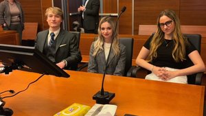 Three young adults sit behind a courtroom desk with a microphone and tissue boxes, while two people stand in the background.