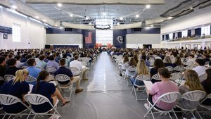 Hundreds of students are seated in a large university gymnasium, attending a formal orientation or convocation ceremony.