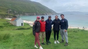 Five people stand arm in arm smiling on green grass near a white building with a rusted roof by a beach and hills.