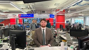 A man in a brown suit and patterned tie sits smiling at a desk in a busy newsroom with multiple monitors and people behind.