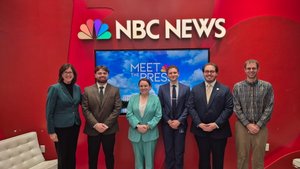 Six people stand side by side in front of a red wall with an NBC News logo and a screen displaying Meet the Press.