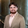 A young man with brown hair and beard smiles wearing a beige blazer and checkered shirt in a softly lit hallway.