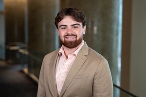 A young man with brown hair and beard smiles wearing a beige blazer and checkered shirt in a softly lit hallway.