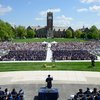 A large outdoor graduation ceremony with hundreds of graduates seated in caps and gowns facing a speaker at a podium.