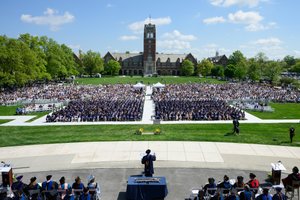 A large outdoor graduation ceremony with hundreds of graduates seated in caps and gowns facing a speaker at a podium.