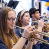 Three people wearing navy shirts play trumpets indoors near blue and yellow balloons.