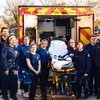 A group of twelve emergency medical responders stand smiling around a stretcher in front of an open ambulance in a suburban.