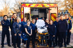 A group of twelve emergency medical responders stand smiling around a stretcher in front of an open ambulance in a suburban.