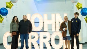 Two men and two young men stand smiling in front of large illuminated letters spelling JOHN CARROLL.