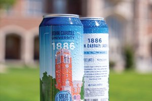 Two cold cans of Great Lakes Brewing Co. 1886 John Carroll Lager sit on a white ledge with a blurred brick building in the