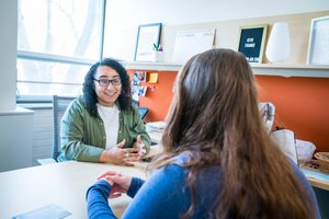 A person with curly hair and glasses talks animatedly across a desk to another person with long hair.