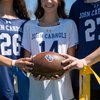 Three people wearing John Carroll sports jerseys hold a football together on a sunny field.