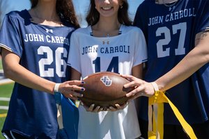 Three people wearing John Carroll sports jerseys hold a football together on a sunny field.