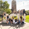 Sixteen happy students celebrate in front of large JCU letters on campus as colorful confetti falls, waving and smiling.