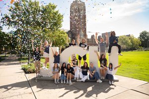 Sixteen happy students celebrate in front of large JCU letters on campus as colorful confetti falls, waving and smiling.