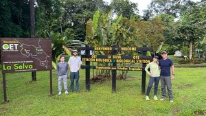 Four people stand smiling on grass beside two large wooden signs welcoming visitors to the La Selva Biological Station.