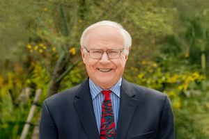 An older man with white hair and glasses smiles while standing outdoors in a dark suit and colorful tie.