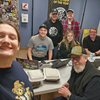 Six people smile toward the camera in a radio station room with laptops and takeout containers on the table.