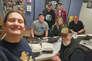 Six people smile toward the camera in a radio station room with laptops and takeout containers on the table.