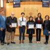 Six people stand in a row indoors, four holding certificates, with banners and a plaque on a wooden wall behind them.