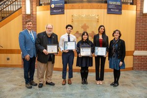 Six people stand in a row indoors, four holding certificates, with banners and a plaque on a wooden wall behind them.