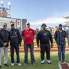 Six men stand side by side on a baseball field with a stadium scoreboard and lights visible behind them.