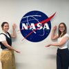 Two smiling students gesture towards a large blue NASA logo on a white wall, celebrating their connection to space.
