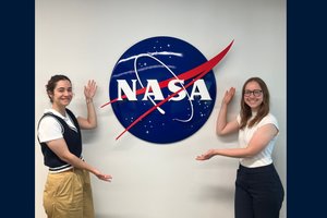 Two smiling students gesture towards a large blue NASA logo on a white wall, celebrating their connection to space.