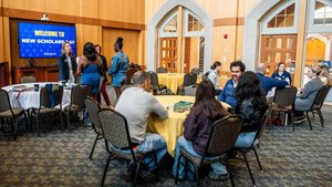 People are sitting and standing in a room with wooden walls and arched windows, with a screen reading New Scholars Day.