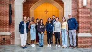 A group of nine people stand smiling in front of a large wooden door with a stone archway on a brick building.