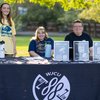 Three people smile behind a table covered with a black cloth displaying the WJCU 88.7 FM logo outdoors on a sunny day.