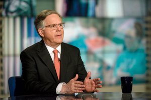A man in a black suit and red tie gestures with his hands while speaking at a table with a dark mug.